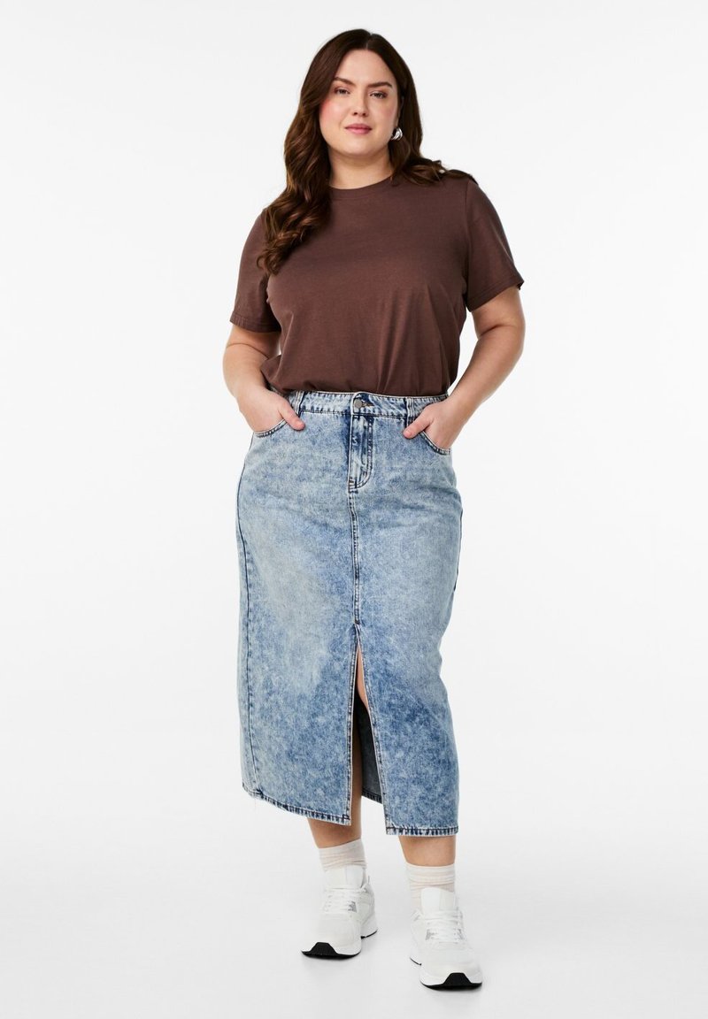 Kvinde iført en brun kortærmet skjorte stukket ned i et lyseblåt denim midinederdel med frontslids, hvide sokker og hvide sneakers, stående med hænderne i lommerne.