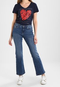 Camiseta navy de escote en V con un diseño de corazón rojo, combinada con jeans de campana en azul claro y zapatillas blancas con detalles en dorado.
