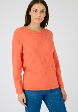 Pull en tricot corail avec un motif en chevrons texturé, des manches longues, un col rond et une silhouette ajustée. Associé à un jean bleu.
