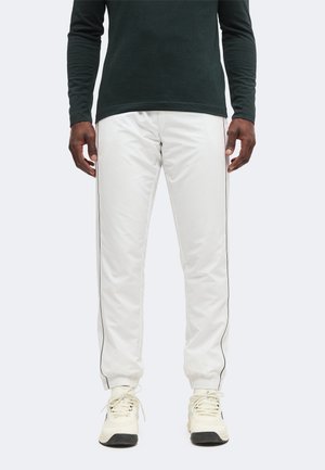 Homme portant un pantalon de jogging blanc avec un liseré noir sur les côtés, un t-shirt noir à manches longues et des baskets blanches sur un fond uni.