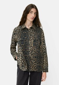 Frau mit langen braunen Haaren, die ein langärmeliges, knöpfbares Oberteil mit Leopardenmuster und schwarze Hosen trägt, steht mit einer Hand in der Tasche.