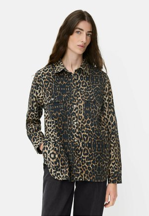 Vrouw met lang bruin haar, gekleed in een lange mouw, knoopjes-overhemd met luipaardprint en zwarte broek, staande met één hand in haar zak.