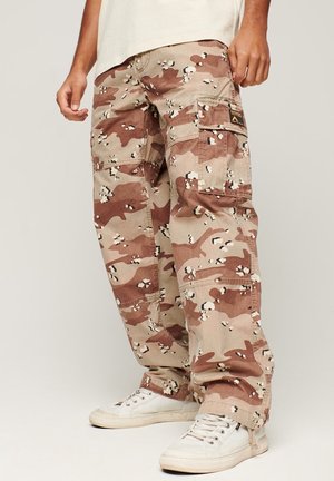 Persoon draagt beige en bruin camouflage cargobroek met meerdere zakken en witte sneakers tegen een effen achtergrond.