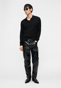 Άνδρας με μαύρο μακρυμάνικο πουκάμισο, μαύρο δερμάτινο παντελόνι, μαύρη ζώνη, μαύρες μπότες και σκουρόχρωμα γυαλιά ηλίου, στέκεται μπροστά σε απλό φόντο.