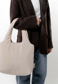 Ljusbeige totebag med en mjuk, vadderad textur, breda remmar och en rundad överkant, hålls av en person i en brun blazer.
