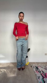 Vrouw die binnenshuis staat, gekleed in een rode top met één schouder, losse lichtblauwe spijkerbroek en bruine puntige schoenen, tegen een effen witte muur als achtergrond.