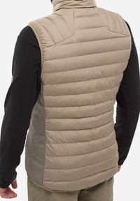 Beige gepolsterte Weste mit aufrechtem Kragen, mit horizontalen Nähten und seitlichen Einsätzen aus einem kontrastierenden Material. Schwarzes Langarmshirt ist darunter sichtbar.