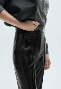 Zwarte nepleer broek met een strak ontwerp, voorzien van een aansluitende snit en een gladde textuur, gecombineerd met een grijze blousecardigan met knopen en een zwarte tas.