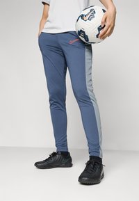 Mørkeblå treningsbukser med lysegrå sidepaneler, myk stoff, tettsittende design, kombinert med svarte sportssko og holder en fotball.