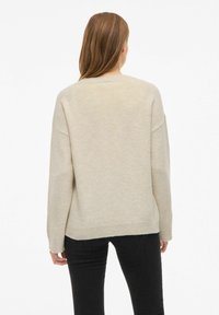 Ženska z dolgimi rjavimi lasmi, oblečena v ohlapen bež pulover in črne hlače, stoji obrnjena stran od kamere proti enobarvni beli ozadju.