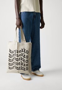 Canvas-Tragetasche mit auffälligem schwarzen "marimekko"-Text in einem wiederholten Muster auf einem natürlichen, beigen Hintergrund; über einem blauen Denim-Outfit getragen.