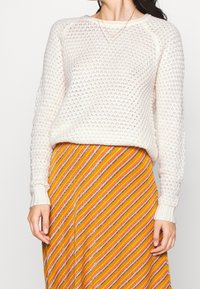 Pull en maille crème avec un motif texturé, associé à une jupe rayée multicolore aux tons orange, rose et gris.