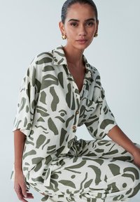 Lehký, volně střižený komplet košile a kalhot v krémově bílé barvě s abstraktními tmavě zelenými vzory. Košile má límec a zapínání na knoflíky.