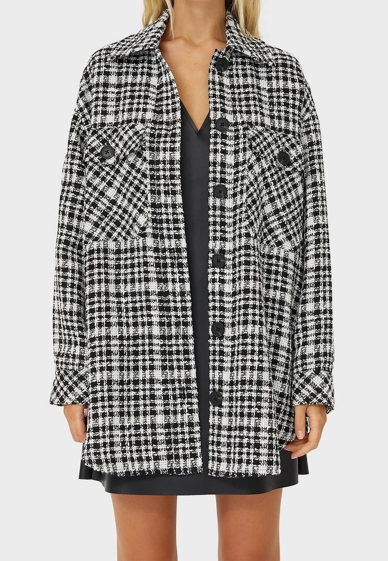 Manteau boutonné à carreaux noir et blanc avec deux poches sur la poitrine porté sur une robe noire à col en V.
