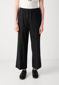 Zwarte wijde broek met een elastische tailleband en trekkoord, met een gladde textuur en minimale accenten. Gecombineerd met zwarte sneakers.