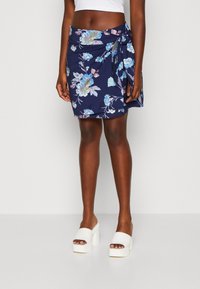 Žena v námořnickém modrém květinovém mini sukně svázaném v pase, bílém crop topu a bílých platformových sandálech na otevřenou špičku, stojící na jednobarevném pozadí.