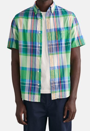 Mand iført en kortærmet skjorte med knapper i grøn, blå og hvid tern over en hvid t-shirt med mørke bukser.