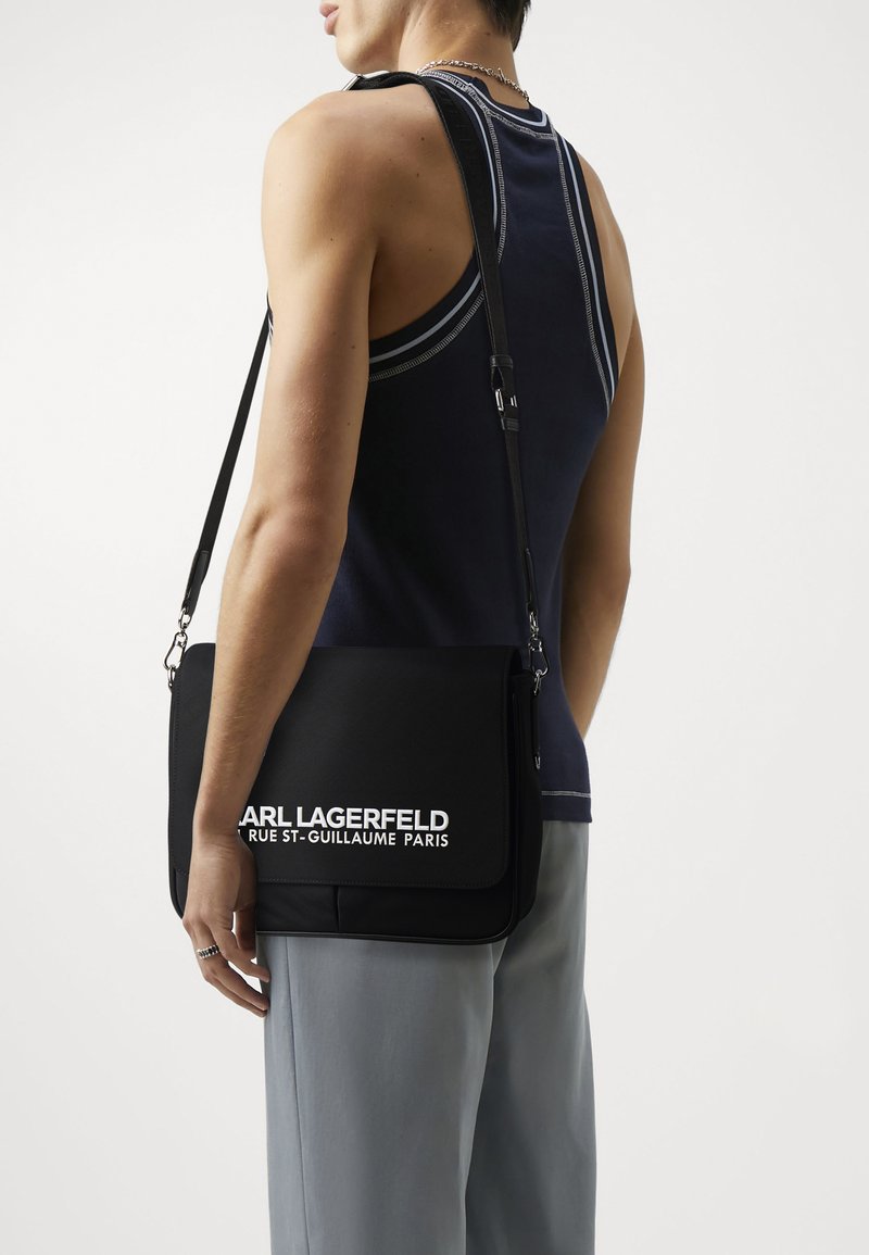 Borsa a tracolla nera in tessuto texturizzato, con tracolla regolabile e un logo bianco ben visibile. Forma rettangolare semplice, design pulito.