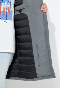 Chaqueta de plumas gris con forro de tejido acolchado negro. Presenta etiquetas de impermeable y cortaviento, cierre de botón y un bolsillo en el pecho del lado izquierdo.