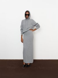 Szürke kötött kapucnis felső és hozzá illő hosszú szoknya szett, kapucnival, bordázott textúrával, fekete hegyes sarkú cipővel párosítva. Napszemüveg is szerepel a szettben.