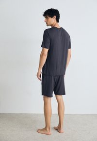 Tricou gri cu mânecă scurtă și pantaloni scurți asortați dintr-un material moale. Design simplu cu o croială relaxată. Modelul este arătat din spate, desculț.