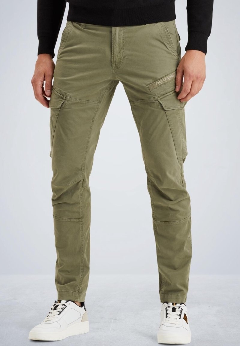 Homme portant un pantalon cargo vert olive avec des poches latérales et des baskets blanches sur un fond uni.