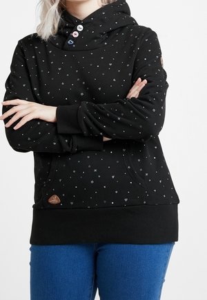 Persoon draagt een zwarte hoodie met kleine hartjes- en stippenpatronen, knoopsluiting, kangoeroezak en blauwe spijkerbroek, armen gekruist.