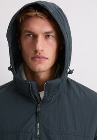 Ciemnoniebieska kurtka z kapturem, z wysokim kołnierzem, przednim zamkiem błyskawicznym i regulowanym sznurkiem przy kapturze, wykonana z gładkiego, teksturowanego materiału.