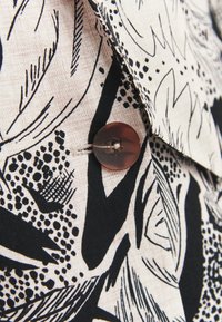 Bouton marron fixé sur un tissu beige avec un imprimé floral abstrait noir et blanc et des motifs linéaires.