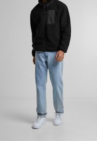 Mężczyzna w czarnej polarowej kurtce, jasnoniebieskich dżinsach i białych tenisówkach stoi na jednolitym szarym tle.