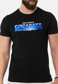 Schwarzes Baumwoll-T-Shirt mit einem blau-weißen Grafikdruck, der "DNM BRAND CIROS BAXX PRIME" über die Brust zeigt.