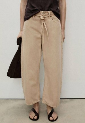 Persoon draagt beige wijde broek met strik aan de voorkant, zwarte sandalen en houdt een donkerbruine tas vast tegen een effen achtergrond.