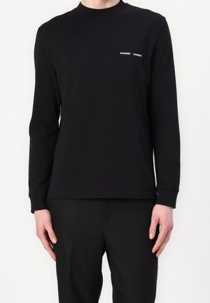 Homme portant un t-shirt noir uni à manches longues avec un petit texte blanc « SAMSØE SAMSØE » sur le côté gauche de la poitrine.