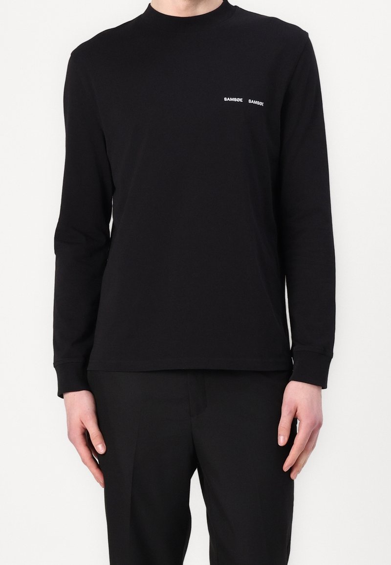 Homme portant un t-shirt noir uni à manches longues avec un petit texte blanc « SAMSØE SAMSØE » sur le côté gauche de la poitrine.