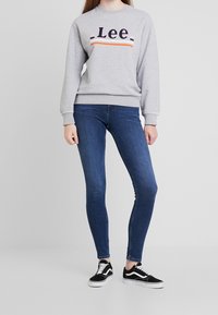 Sudadera gris con el logo "Lee" y rayas de color. Jeans ajustados de mezclilla oscura y zapatillas negras con suelas y cordones blancos.