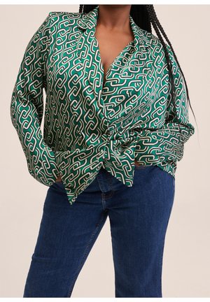 Vrouw draagt een groen patroon blouse met lange mouwen en een geometrisch kettingontwerp, gecombineerd met een blauwe spijkerbroek, staand met handen in de zakken.