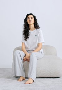 Camiseta blanca de algodón con logo verde, combinada con pantalones anchos de color gris claro. Sentado en una silla lounge texturizada de color claro.