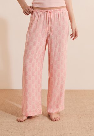 Personne portant un pantalon de pyjama rose ample avec un petit motif ananas, debout pieds nus sur une surface texturée.