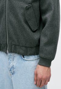 Grijze zip-up jack van gestructureerde stof, met een voorzak en ribgebreide manchetten, gecombineerd met lichtblauwe denim jeans.