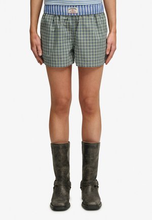 Nogi w zielono-niebieskie kratkę szorty z niebieskim paskiem w pasie i ciemnoszare, skórzane buty do połowy łydki z paskami.