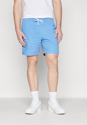 Homme portant un short bleu clair à cordon, un t-shirt blanc, des chaussettes blanches montantes et des baskets blanches, debout devant un fond uni.