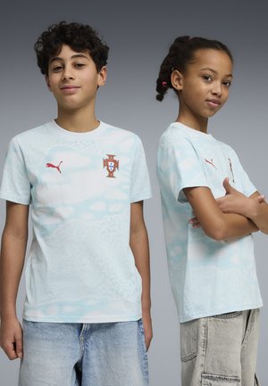 Dos niños vistiendo camisetas de fútbol de Portugal en azul claro con logos de Puma, uno mirando hacia adelante y el otro con los brazos cruzados, sobre un fondo gris.