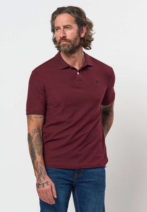 Homme avec barbe et tatouages portant un polo bordeaux et un jean bleu, se tenant devant un fond clair uni.
