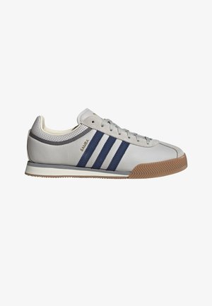 Sneaker Adidas Samba bianco con strisce blu navy, suola in gomma, lacci grigi e collarino traforato sul tallone, mostrato dal lato esterno.