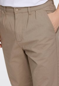 Beiga byxor i texturerat tyg, med knappstängning och sidofickor, som visar veck för extra designdetalj.