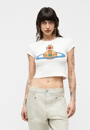 Mujer con cabello oscuro y tatuajes lleva una camiseta blanca recortada con diseño colorido de planetas y pantalones blanco crema contra un fondo liso.