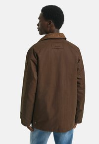 Bruine jas met een corduroy kraag, voorzien van een label op de rug, drukknoopjes op de mouwen en een comfortabele pasvorm. Zichtbare stiksels en een zachte textuur.
