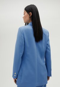 Blazer azzurro chiaro con spalle strutturate, spacco singolo sul retro e chiusura a tre bottoni sulle maniche. Tessuto liscio con texture minimale.