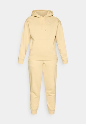 Beige Puma-Hoodie und Jogginganzug, aus weichem Material gefertigt. Mit einer Kängurutasche, gerippten Ärmeln und Logo-Akzent auf Brust und Oberschenkel.