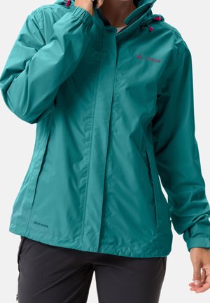 Person iført teal vandtæt jakke med hætte og sorte bukser, justerer krave med den ene hånd.